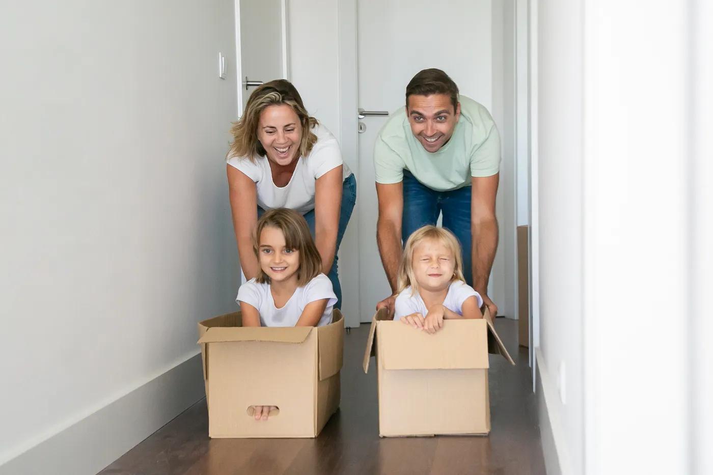 Familia riendo mientras empuja a sus hijas dentro de cajas de mudanza en el pasillo de su hogar
